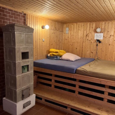 Ein beigefarbener mit Kacheln bestückter Schwedenofen ist Blickfang im Ruheraum, in dem große, erhöht gelagerte, bunte Matratzen die Möglichkeit zum Entspannen bieten.