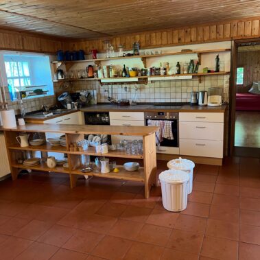 Die Gruppenküche ist der Ort des gemeinsamen, miteinander Kochens. Sie ist L-förmig und wird durch ein niederes Abstellregal, das in den Raum ragt, zum U. Ein Gewürzregal an der Wand komplettiert die große Küche.